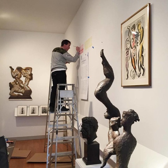 Photo of a white gallery with light brown wood floors and a young man standing on a metal ladder painting the wall to the right of us. On the wall to the left behind him there is a sculpture hung of two female figures, one laying down with the other balancing on her legs. To the right front of him there are three dark brown sculptures of people, and the illustration by Marc Chagall, &quot;The Circus Performers&quot;.