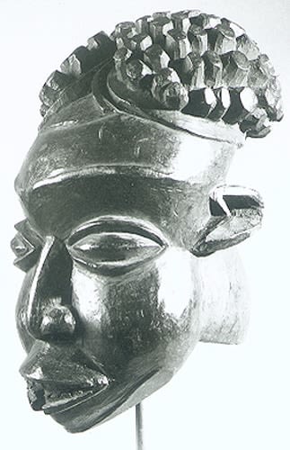 Stained wooden mask of an individual, its face elongated, facial features enlarged. It wears a headdress in form of a chief's knitted cap which parts its hair in two.