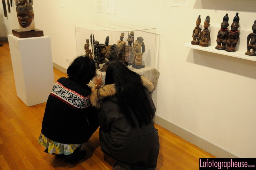 Two adults with dark hair wearing winter clothing are crouched to look at a collection of small sculptures in a glass box on a white, rectangular mount. To the left of them is a wood sculpture of a human face, and to the right of them are a series of twin sculptures standing on a wall mounted shelf.