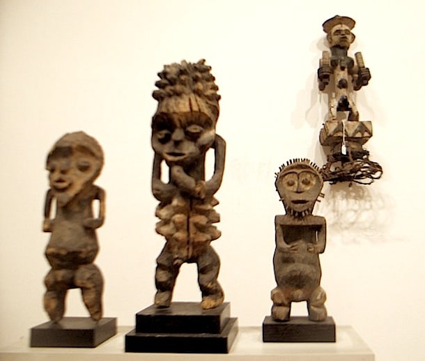 Four wood sculptures of humanoid, geometric shapes face us directly. Three of them are standing on a white block mount, and another is mounted on the white wall to the left.