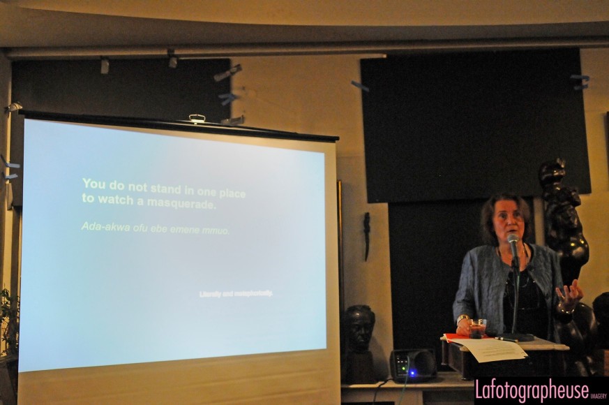 Adult in a blue cardigan over a black dress stands to the right of us at a podium, speaking into a microphone. To the left is projector with a blue image with white text on it, &quot;You do not stand in one place to watch a masquerade.&quot;