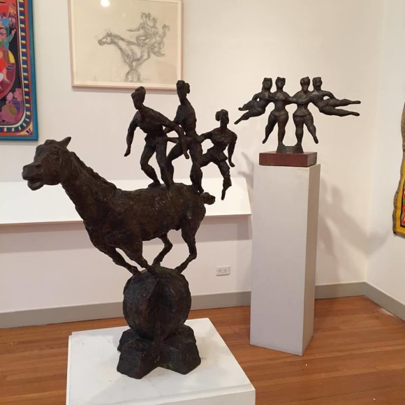 Photo of a white gallery wall with light brown wood flooring beneath it. In front of us is a large black sculpture of three tiny people balancing on a gigantic horse. Behind this sculpture is another black sculpture by Chaim Gross, &quot;The Flying Ballerinas&quot; which is a symmetrical composition of four female figures, one standing and holding the other as they fly horizontally outwards on each side. On the wall behind the sculptures is a large line drawing of the horse sculpture in a light cream frame. Next to the drawing the corner of a large, colorful artwork is barely visible.