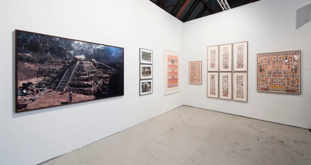 Installation view of an art fair booth, showcasing paintings, collages, and photos on the walls.