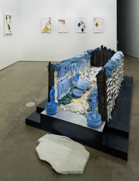 Mixed media pieces line the walls and a large rectangular installation is on the floor.