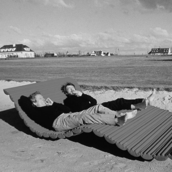 Archival photo of Pierre Paulin and &Eacute;tienne Fermigier laying beachside on a lounger smoking cigarettes