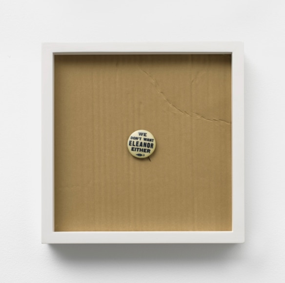 Photograph of a pin that says "we don't want Eleanor either" on top of a material that resembles cardboard in a white frame