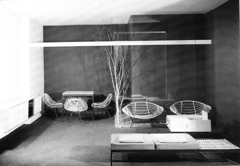 Foreground seating area features a bench and rattan chairs while the background seating area features rattan chairs around a dining table at a&nbsp;furniture fair in 1957