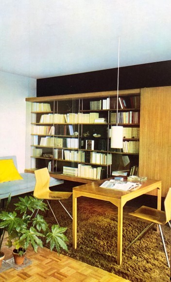 Archival photo of a desk atop a shag carpet and a wooden and glass bookcase in the background.