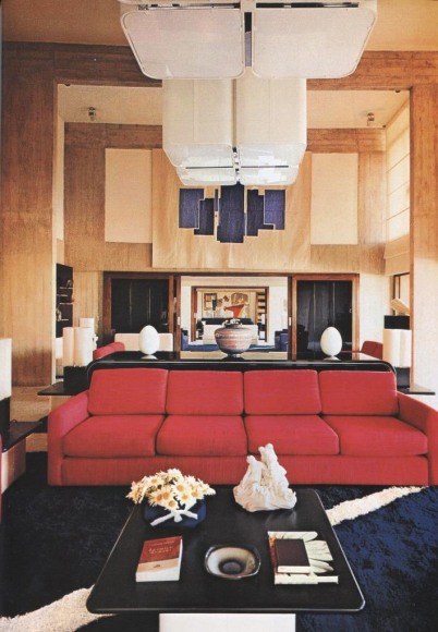 Archival photo of a living room with high ceilings, focal ceiling lights, a red couch, and a white and black coffee table in the foreground