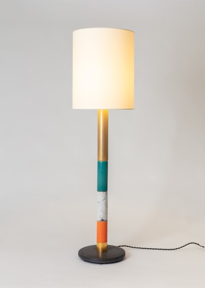 Floor lamp in various colors, white fabric shade. Colors from bottom to top, black base, gold section, orange section, white, green raku, gold. Light turned on.