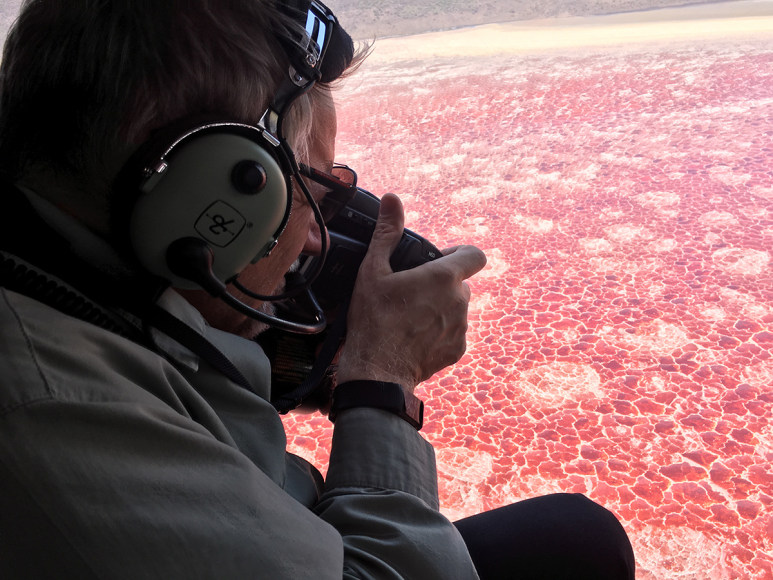 Portrait of Photographer Edward Burtynsky Shooting Lake Natron, Tanzania, 2017. He is in an aircraft, hanging out the door, shooting the bright pink lake.