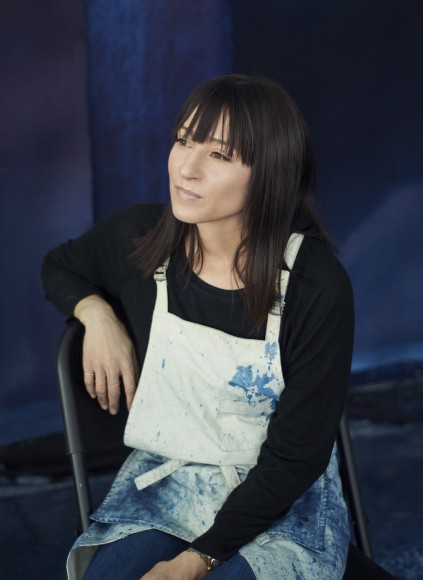 Portrait of Artist Miya Ando wearing a painting apron, seated in front of an indigo painting.