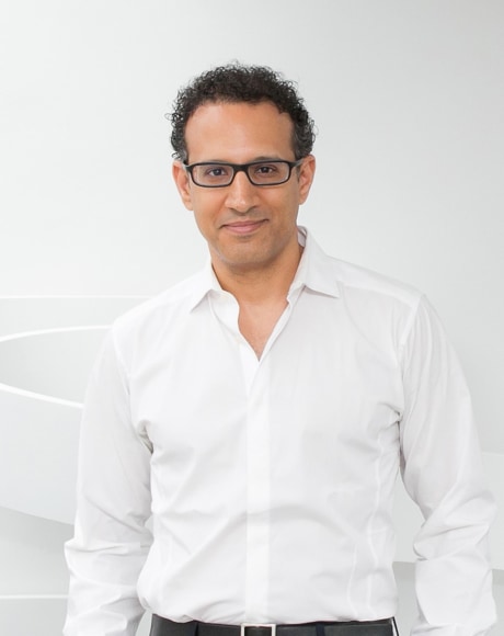 A portrait of artist Ghiora Aharoni wearing a white button down shirt in front of a white sculpture.