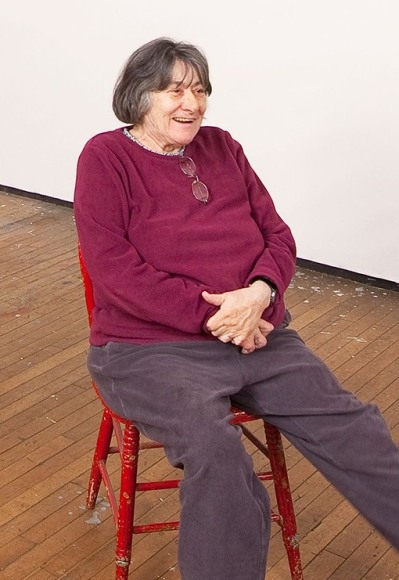 Portrait of Artist Susan Weil in Her Brooklyn, New York, Studio, casually Seated in a red chair, wearing a purple sweater.