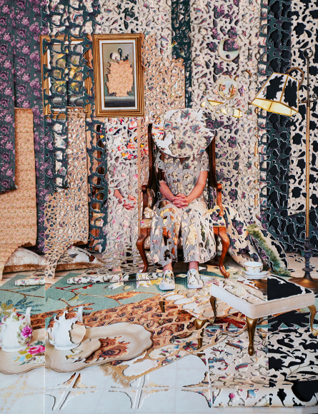 A woman sits in a patterned room with a hat concealing her face. Her clothes match the wall paper which lets her blend into the background