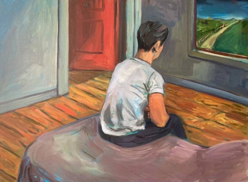 A young man sits in a room, on a bed with his back to the view, looking out the window at a road in the distance