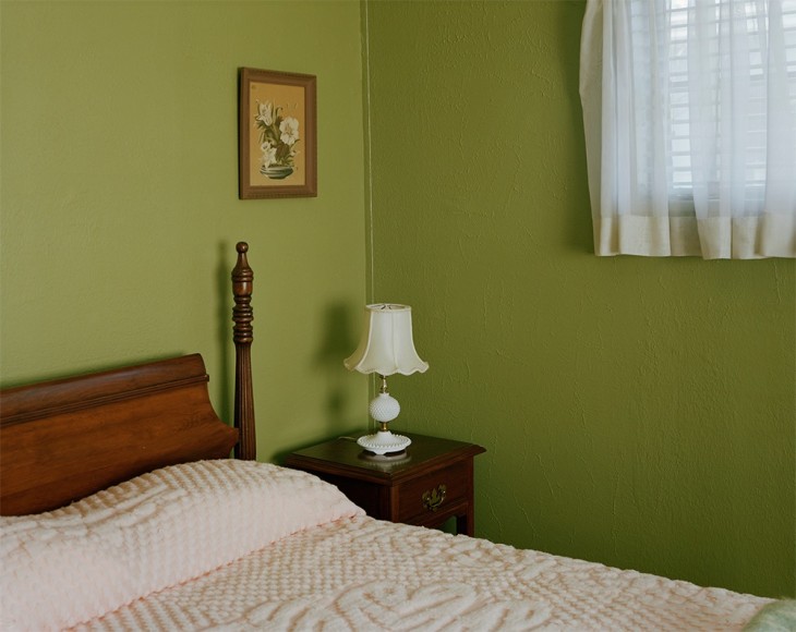 Horizontal photograph of simple bedroom with green wall, sheer curtains and a simple lace coverlet on the bed. There is a nightstand with one lamp to the right and a botanical print on the wall above