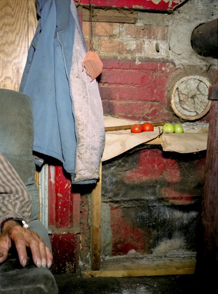 ike Smith (1951-)  Carter County, TN (05-191-14R), 2005  Archival Pigment Print  40h x 30w in 101.60h x 76.20w cm  MS_017, color photograph of the interior of a cabin, a man's hand rest n the arm of a chair to the left, towels hang behind him, green and red tomatoes are lined up on a shelf on the wall.