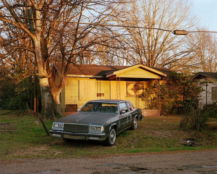 Horizontal photograph of a yellow ranch style house with an early model car in the driveway facing the viewer
