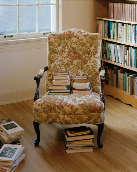 Vertical photograph of a upper middle class living room, with a floral covered wind chair with a stack of books on the seat and full bookclases on the
