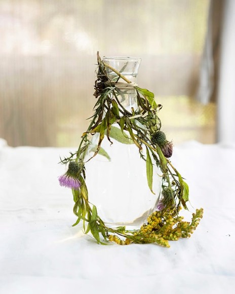 Color photograph of leaves and flowers wrapped around glass vase on table, by James Henkel