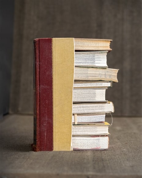 James Henkel  Chapters From The Series Books, 2020  20h x 16w in 50.80h x 40.64w cm  Edition 5  JHe_055 a bottom view of 9 books ranging from 100-250 pages each stacked on top of each other with a book cover that has been partially cut partially covering the stack of books