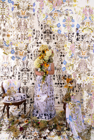 A woman in a floral printed dress stands in the middle of a room with her face concealed by a bouquet of flowers. The surrounding floor, walls and furniture are all covered with the same pattern as her dress