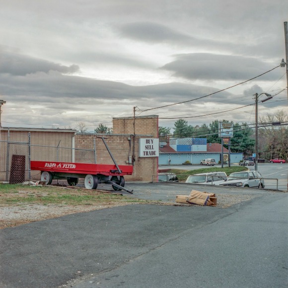 Ken Abbott  Radio Flyer and Pawn Shop, Patton Ave, November 2004, Printed 2020  Archival Pigment Print  Image Size: 7 x 7 in Paper Size: 12 x 9 1/2 in Mat Size: 14 x 11 in  Edition of 10  $ 300.00 unframed, photograph of a large red wagon in front of a grey warehouse