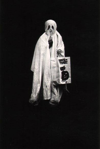 21. Shawn Walker, Halloween, Brooklyn, ​1960s. Figure in a draping white ghost costume faces the camera holding a trick-or-treat bag. Background is completely black.