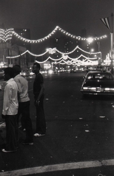 17. Anthony Barboza, Coney Island, NY, ​1970s. Three figures gathered on the left of the frame. Cars and string lights are out of focus in the street behind them.