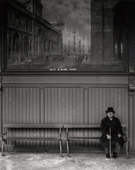Nino Migliori, Northern People, 1950. A man with a cane seated on a bench. A mural of Milan is on the wall behind him.