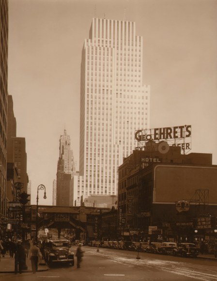 Paul J. Woolf, Untitled, c. 1933. Daytime street scene featuring a building marked &quot;Geo. Ehret's&quot; in the foreground right on a street lined with cars. A tall building looms in the background center.