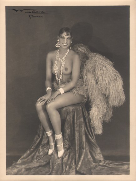 Lucien Walery, Josephine Baker, c. 1925. Subject poses seated, topless, in large jewelry and feathers.