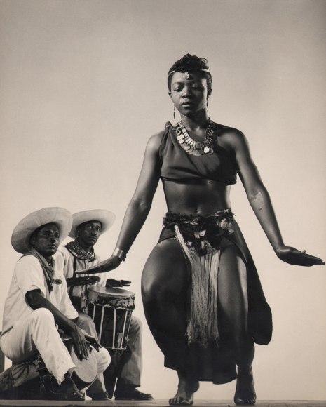 Gjon Mili, Pearl Primus, ​c. 1943. Subject crouches with eyes closed to the right of the foreground, while two male musicians look to her while playing drums in the background.