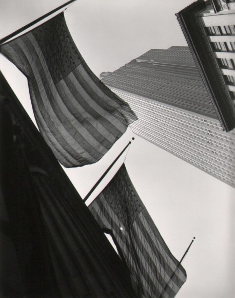 27. John C. Hatlem, Empire State Building, ​c. 1935. Street-level view looking up at the base of the Empire State Building, The view is partially obscured by two American flags hanging from the building nearest the photographer.