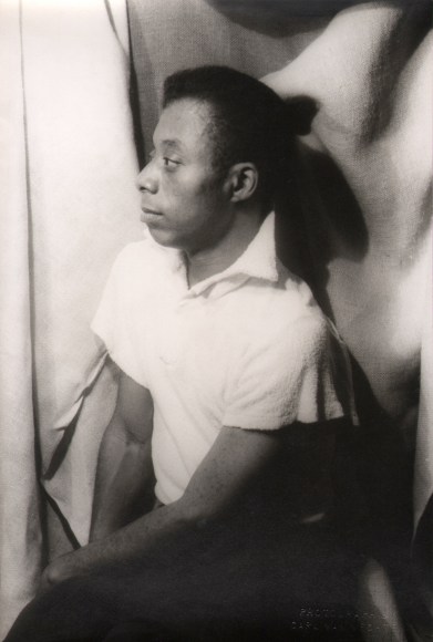 29. Carl Van Vechten, James Baldwin, ​1955. Seated portrait against a draping fabric backdrop with subject facing the left of the frame.