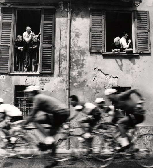Stanislao Farri, Passa il Giro, ​1956. A group of cyclists, blurred with motion, pass a building with onlookers observing from two open-shuttered windows.