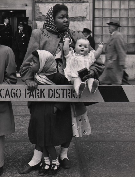 Marvin E. Newman, Mother, Daughter, &amp; Doll Await the MacArthur Motorcade, 1951. A woman holding a large baby doll and young girl, both in headscarves, stand behind a barricade. Men walking by and police officers can be seen behind them.