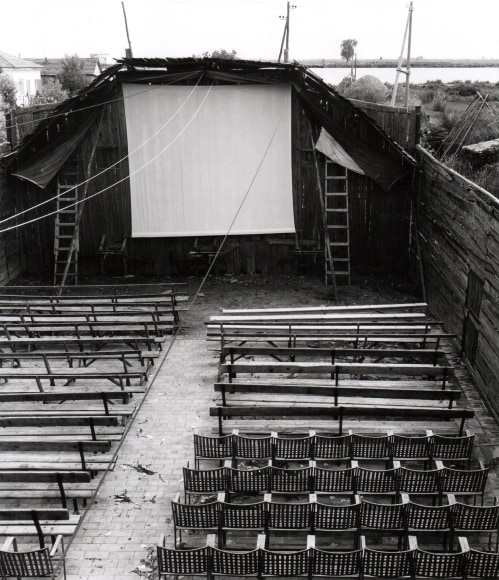Pietro Donzelli, Cinema in Pila, Veneto, ​1954. Outdoor movie screen with rows of seats and benches in front of it. Ladders are propped up on either side of the screen.