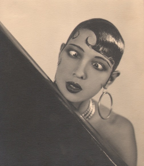 George Hoyningen-Huene, Josephine Baker, ​c. 1929. Subject crosses her eyes and wears large hoop earrings, resting her chin on a black piece of furniture.