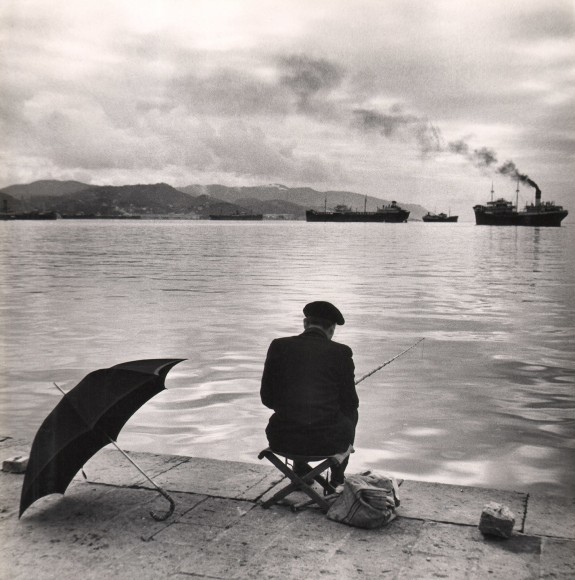 Ezio Quiresi, Domenica nel Porto, ​1956. Seascape with a man in black fishing, back to the camera, in the foreground. Steam boats and mountains are visible in the distance.