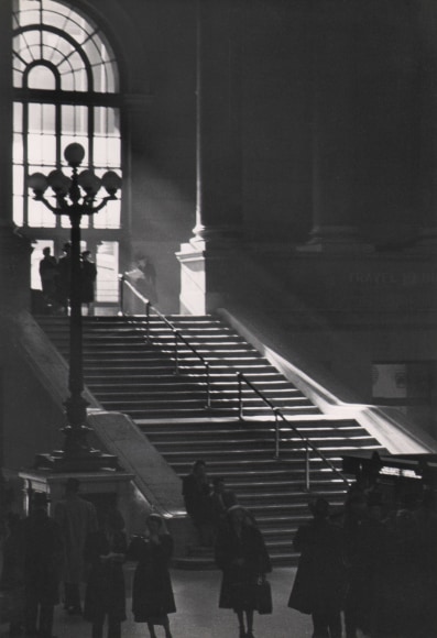 35. Fritz Neugass, Penn Station, ​c. 1948. Light streams into the station diagonally down a set of stairs from a large window on the top left of the frame. Silhouettes of patrons are at the bottom of the stairs and frame.