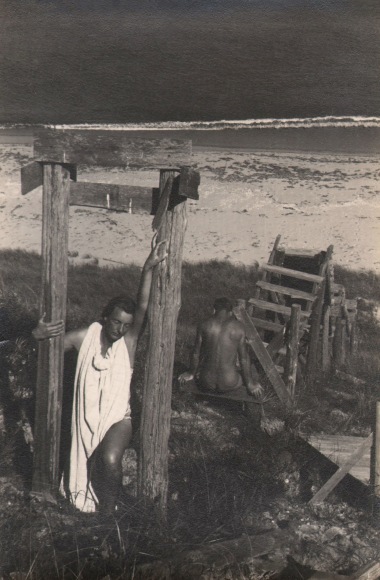 PaJaMa, Margaret French &amp; Jared French, Nantucket, ​c. 1945. A woman in draping white fabric poses between two wooden posts. A nude male figure is seated, back to the camera, in the midground. The beach and sea are in the background.