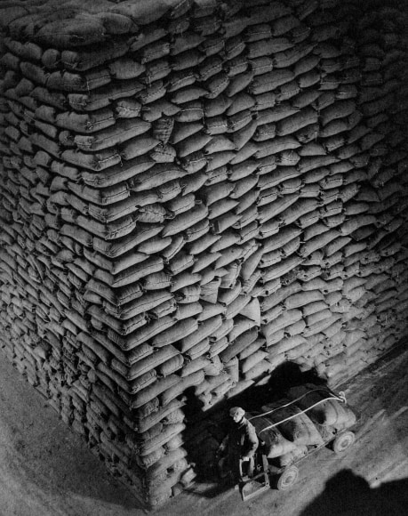 Harold Haliday Costain, Untitled (Sugar Sacks), ​1935. A man in the lower right of the frame pulls a cart in front of stacked sugar sacks.