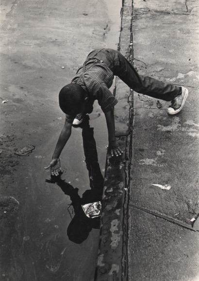 29. Beuford Smith, Bed-Stuy, Brooklyn, ​1970. A boy reaches into a puddle where the sidewalk meets the street.