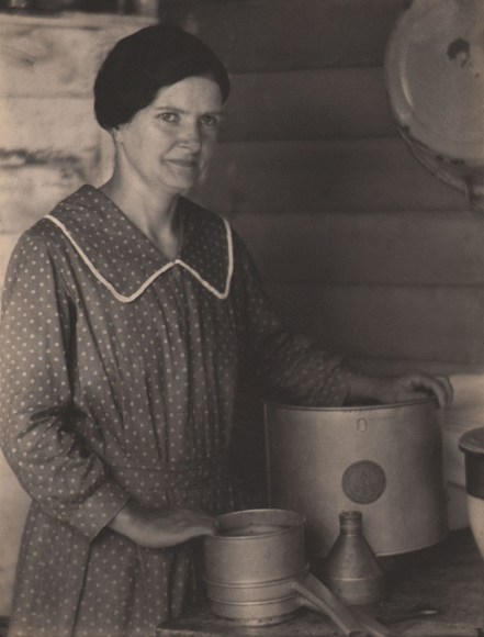 Doris Ulmann, Untitled, ​1928&ndash;1934. Woman with dark hair in a polka-dot dress standing at a table with hands resting on metal containers.