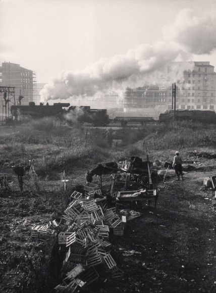 Mario de Biasi, Outskirts of Milan, Ortica, ​1949. A pile of wooden crates is seen before a horse-drawn cart with a man standing beside it. A train emitting smoke clouds divides the frame horizontally, some buildings can be seen behind it.