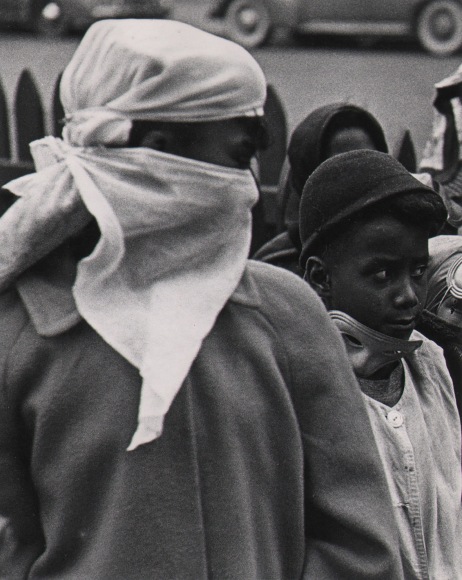 Marvin E. Newman, Chicago, ​1950. Close-up on two figures in a crowd with heads turned to the right of the frame. One wears a headscarf and bandana around his lower face, the other is in a hat with a masquerade mask around their neck.