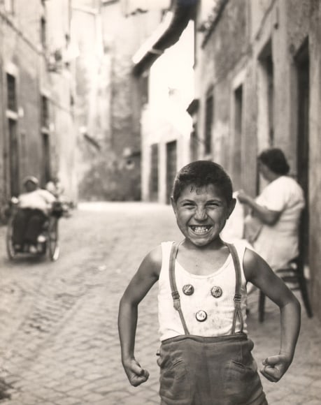 Carlo Bavagnoli, Gente di Trastavere, 1957&ndash;1958. A young boy poses in an alleyway, flexing his muscles and smiling to the camera. He wears suspenders and three bottle cap buttons on his shirt.