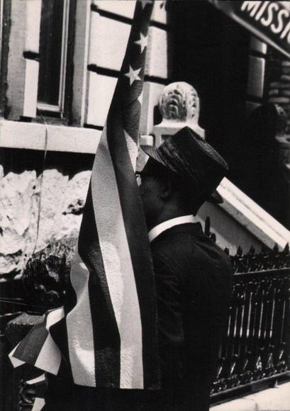 22. Shawn Walker, Untitled, ​c. 1965. Man in a jacket and hat carries an American flag, facing away from the photographer.